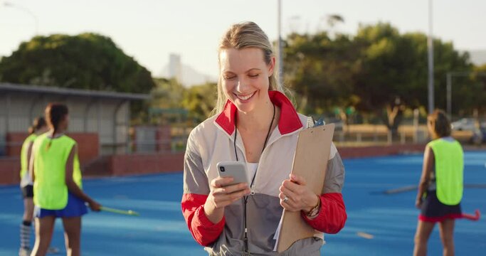 Smiling hockey coach texting on phone, searching and browsing internet while standing on sports pitch. Young happy trainer using technology to monitor team progress. Confident woman holding clipboard