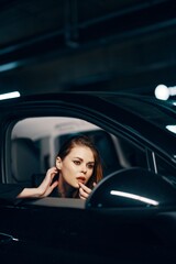 Obraz na płótnie Canvas a vertical photo from the side, at night, of a woman sitting in a black car and looking out of the window and gently touching her face while adjusting her makeup looks into the side view mirror