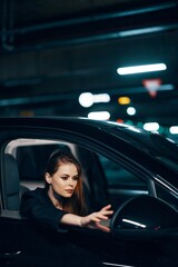 vertical photo from the side, at night, of a woman sitting in a black car and looking out of the window and reaching out to the side view mirror to correct it