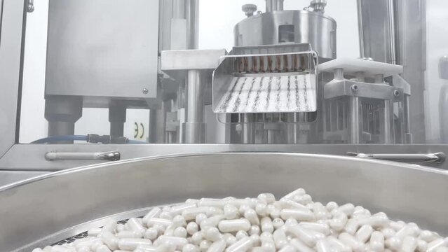 White capsules pills moving on metal automatic line in workshop of pharmaceutical factory. Closeup view of drugs move along conveyor belt during work process in pharma chemical company. 