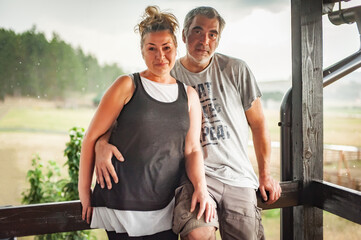 Happy embracing couple, Generation X, posing, leaning against a fence, in rainy weather on the beautiful green hills of the mountain