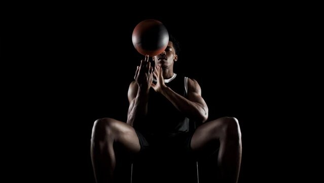 Basketball player spinning a ball. African american sports man sitting and practicing against black background.
