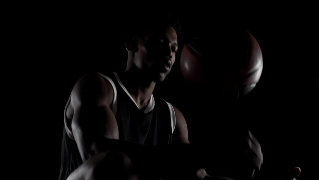 Basketball player sitting on a bench. African american man balancing a ball against black background.