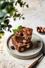 Stack of chocolate brownies with hazelnuts and mint leaf on a plate and greenery in blur