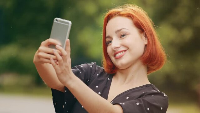 Portrait stylish beautiful girl caucasian attractive redhead woman blogger female model stands outside in park holds smartphone takes selfie photo on mobile camera records vlog filming video for blog