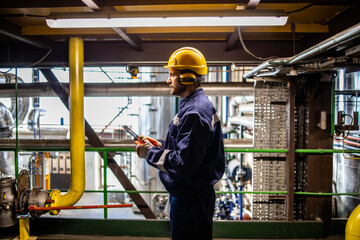 Professional factory worker wearing yellow hardhat and earmuffs standing in petrochemical refinery looking at his computer.