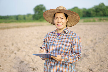  Asian woman farmer is at agriculture land, wear hat, plaid shirt, holds and write on paper clipboard. Concept : research and analyze soil and environment to improve  and solve  agriculture problems. 