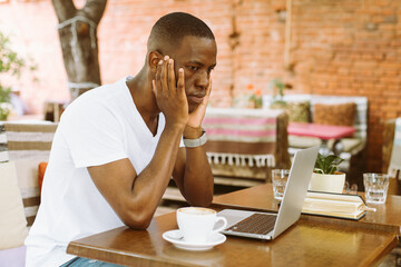 Concentrated and serious african afro american businessman using laptop in cafe with head in hands in outdoor cafe. Video calling and reading finance information. Business and freelancing problems