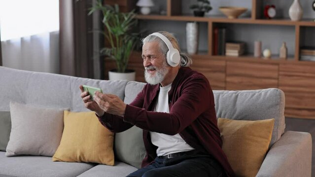 Happy handsome elderly gray-haired man in white headphones holding smartphone and laughing. Old man sitting on couch at home watching funny content videos blogs, TV shows, films.