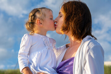 Portrait of a little girl and her mother. Mom and daughter are cute hugging against the blue sky with clouds. Mom and daughter in white clothes, family happiness.