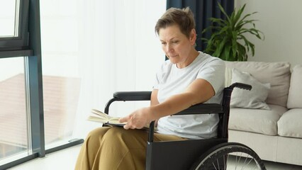 Senior woman sitting on the wheelchair reading a book at home. Leisure at home, hobby