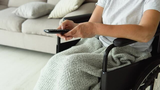 Senior lady in wheelchair watching tv in living room holding remote control changing tv channels. Grandma turning on the TV