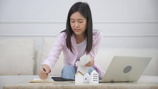 Young asian woman sitting and planning and calculate expense and mortgage with calculator and home on desk, insurance and budget of residential, loan and residence, business and property concept.