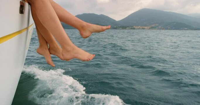 Two carefree friends on tropical vacation break, swinging, dangling legs and feet from yacht. Closeup of relaxing women enjoying a ocean boat cruise in Italy. Tourism abroad, travel overseas in summer