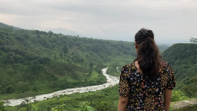 Rear view of young Indian girl watching the mountain and river stream at hill top in India at sunset.