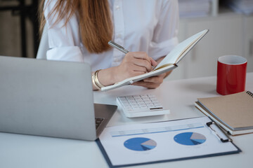 Close up of business hands are writing on the notebook with coffee cup and paperwork shown chart on the table.