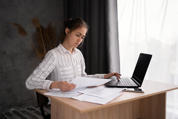 hispanic young business woman working with documents using laptop in home office, analyzing work results, doing paperwork at workplace using computer software
