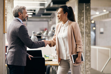 Asian businesswoman handshaking with mature businessman at corporate office. - obrazy, fototapety, plakaty