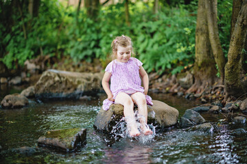 Cute little toddler girl having fun by a river on warm and sunny summer day. Happy excited preschool child splashing with water in forest stream creek.