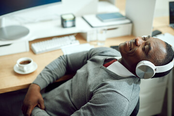 Smiling black businessman with eyes closed enjoys in music over headphones in office.