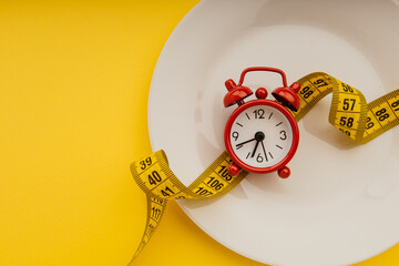 Red alarm clock on white plate with measuring tape on a yellow background close-up