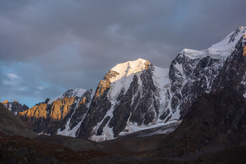 Gloomy landscape with giant snowy mountain top and sunlit gold rocks in dramatic gray sky. Huge...