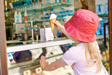 Cute little toddler girl choosing and buying ice cream in outdoor stand cafe. Happy preschool child with glasses looking at different sorts of icecream. Sweet summer dessert