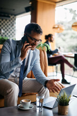 Happy businessman using laptop while communicating on cell phone in cafe.