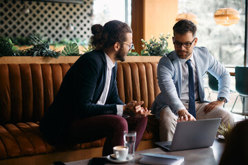 Young businessman and his colleague talk while using laptop during meeting in cafe