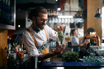 Young bartender preparing mojito cocktail and adding mint leaf in glass at bar counter. - obrazy, fototapety, plakaty