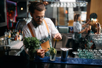 Young barmen adding mint leaf while preparing mojito cocktail at bar counter.
