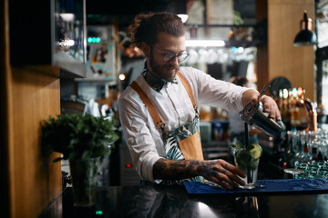 Happy bartender pouring cocktail from a shaker at bar counter.