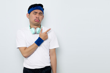 Portrait of sad young Asian sportman in blue headband and sportswear white t-shirt with headphones, pointing fingers at copy space, advertisement banner isolated on white background
