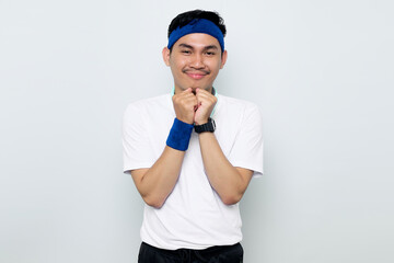 Smiling young Asian sportman in blue headband and white t-shirt with headphones, keeps hands under chin, look at camera, remembers pleasant moment isolated on white background. Workout sport concept