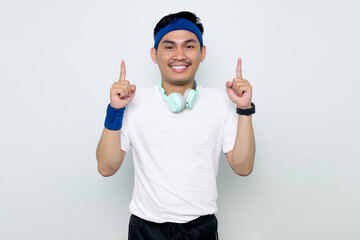 Fototapeta premium Excited young Asian sportman in blue headband and sportswear white t-shirt with headphones, pointing fingers up having a good idea isolated on white background. Workout sport concept