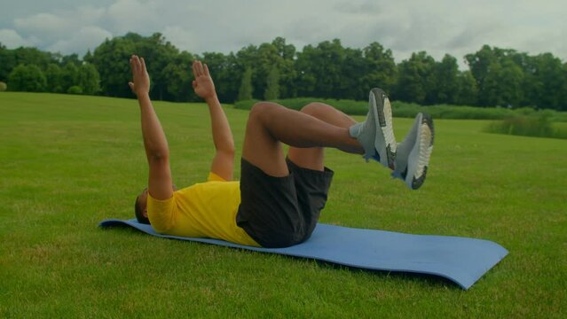 Determined active sporty fit African American man lying on fitness mat, practicing dead bug exercise, strengthening and stabilizing core, spine, and back muscles while training in public park.