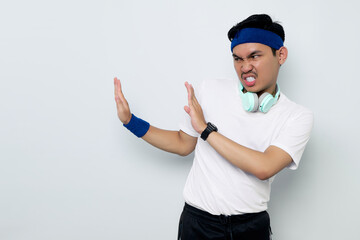 serious young Asian sportman in blue headband and sportswear white t-shirt with headphones, showing stop gesture, demonstrating denial sign, rejecting something unwanted isolated on white background