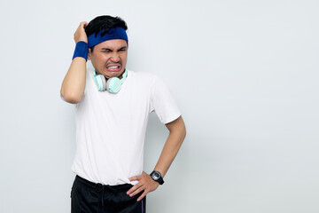 Stressed young Asian sportman in blue headband and sportswear white t-shirt with headphones, touching his temples and having headache isolated on white background. Workout sport concept