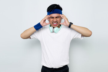 Stressed young Asian sportman in blue headband and sportswear white t-shirt with headphones, touching his temples and having headache isolated on white background. Workout sport concept