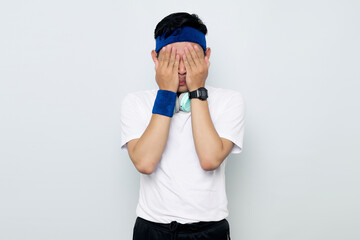 Portrait of young Asian sportman in blue headband and sportswear white t-shirt with headphones, covering face with hands isolated on white background. Workout sport concept