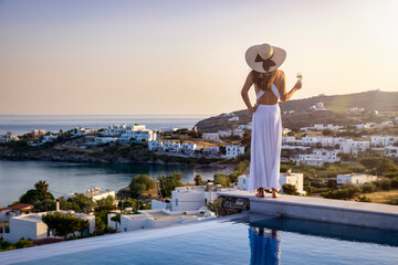 A elegant woman in a white dress stands by the pool with a drink and enjoys the view over the...