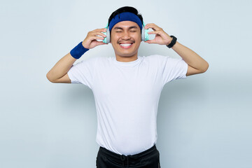 Naklejka premium Portrait of cheerful young Asian sportman in blue headband and sportswear white t-shirt while listening music favorite with headphones isolated on white background. Workout sport concept