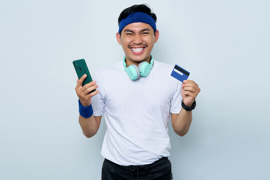 Portrait of cheerful young Asian sportman in blue headband and sportswear white t-shirt while listening music with headphones, holding mobile phone and credit card isolated on white background