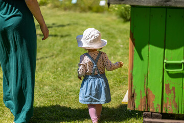 mother in green dress and daughter in denim dress and white hat walk together on grass at sunny day, green abandoned bee hive - obrazy, fototapety, plakaty
