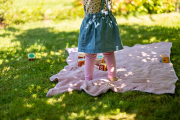 baby girl in denim dress have fun outdoor, sunny day and grass, toddler stand barefoot on the picnic blanket with wooden toy blocks, little legs