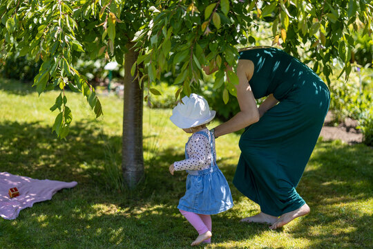 mother in green dress and daughter in denim dress walk together barefoot under the tree, outdoor place for toddler to have fun at sunny summer day in the shade