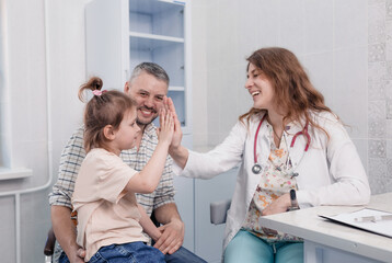 a young doctor girl gives five to a cute little patient for a consultation with her dad in the hospital. A smiling pediatrician cheer up a small child in the clinic.