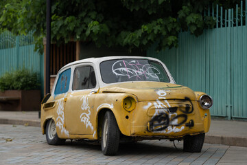 Damaged retro old yellow automobile parked at sidewalk in city with green metal wall and plants in background. Vintage dirty rusty car in poor condition. Forgotten unnecessary things, useless stuff.
