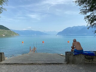 Mann sitzt mit blauer Badehose mit dem Rücken zu uns auf Mauer und hat gigantischer Aussicht - See, türkis, Berge, Himmel, blau