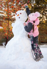 Little girl dressed in winter clothes making hair with twigs for snowman in park in daytime while...